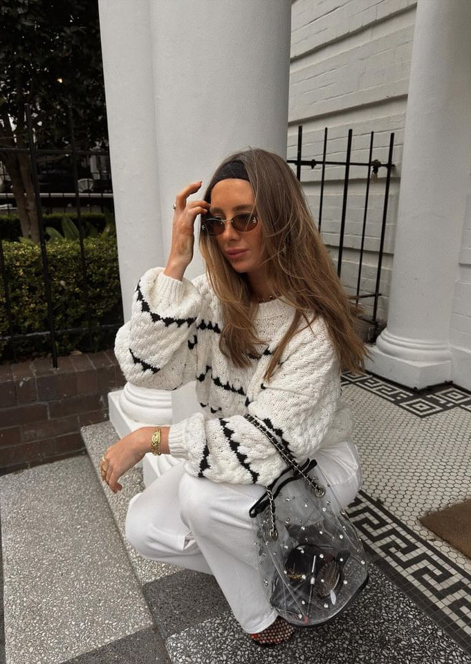 Cream chunky knit sweater with black heart pattern, worn with white pants, front view with hand on head