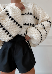 Cream chunky knit sweater with black heart pattern, worn with black shorts, front view with hand adjusting sweater