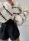 Cream chunky knit sweater with black heart pattern, worn with black shorts, front view with hand adjusting sweater