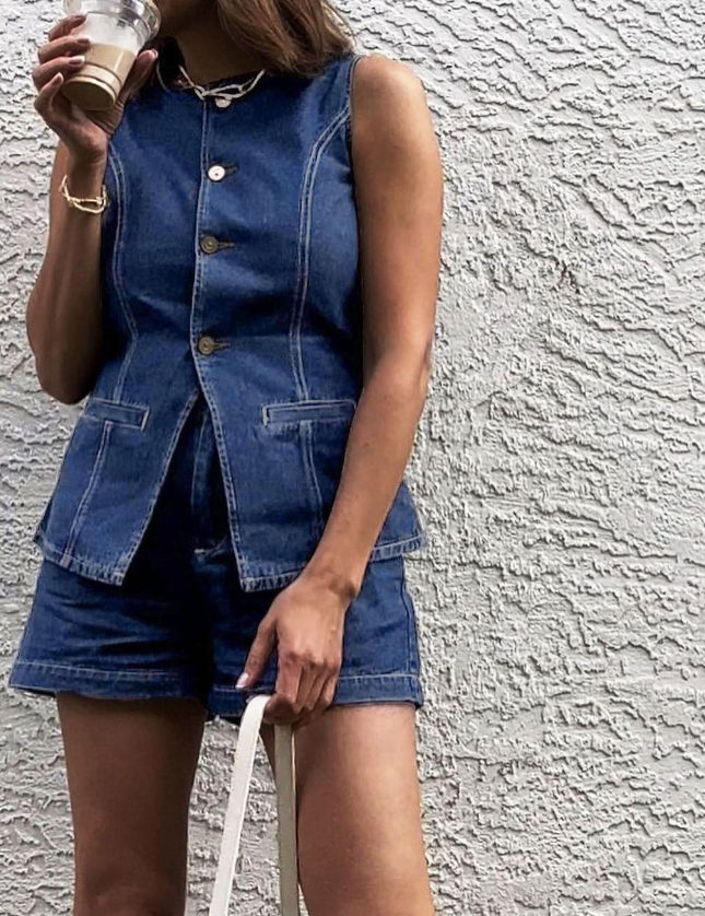 Woman wearing denim playsuit with gold buttons and pockets, holding coffee cup