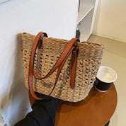 Woven straw tote bag with brown leather handles, side view on table with cup