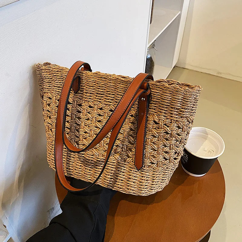 Woven straw tote bag with brown leather handles, side view on table with cup